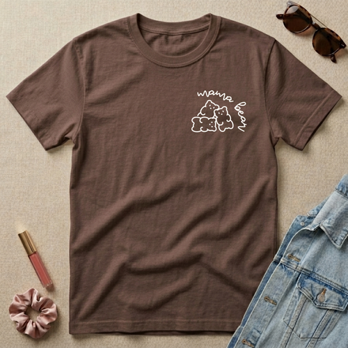 Brown t-shirt with 'mama bear' graphic on a beige surface with sunglasses, scrunchie, and lip gloss.