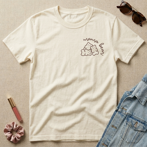 Beige t-shirt with 'mama bear' graphic, sunglasses, scrunchie, and lip gloss on a beige surface